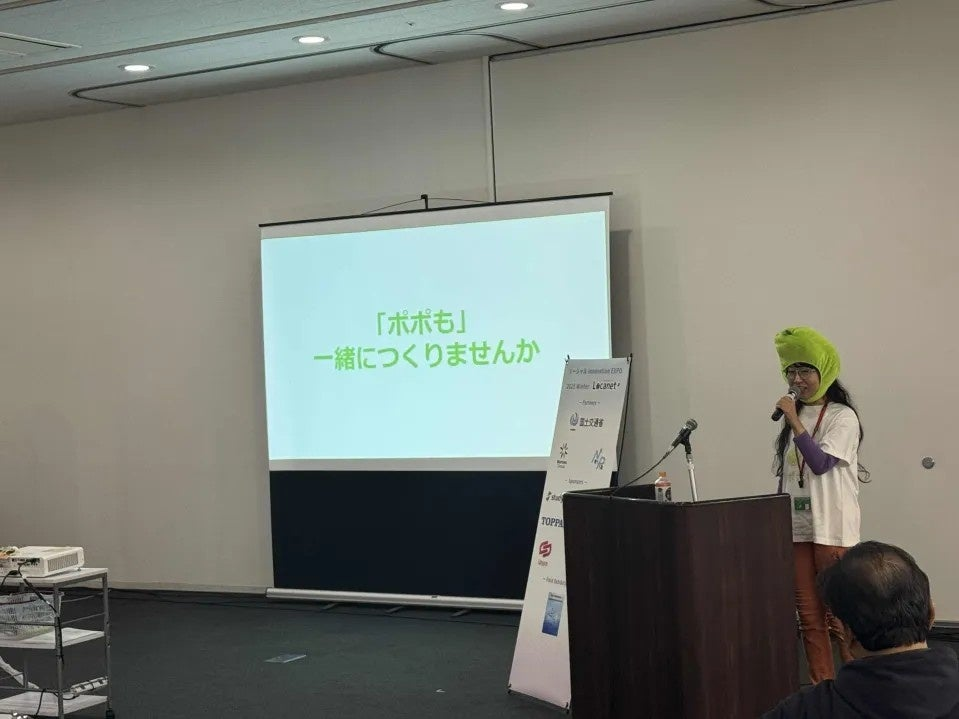 Sabii Ishida speaks at a lectern beside a projected Popomo presentation in Japan